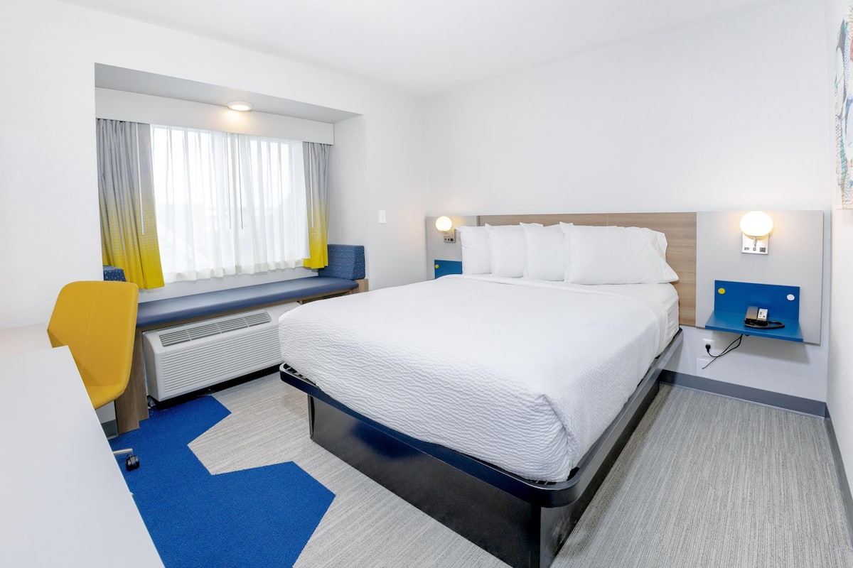 A hotel room with a bed, a desk, a chair, a window with curtains, and a wall-mounted nightstand.