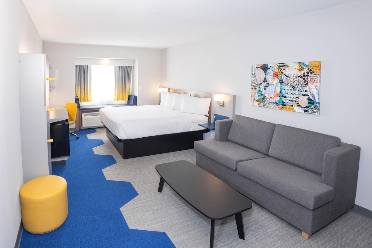 A hotel room with a bed, a grey sofa, a small table, a yellow stool, and a colorful abstract painting on the wall.