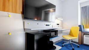 Une chambre d’hôtel moderne équipée d’un téléviseur à écran plat, d’un bureau avec un ordinateur, d’une chaise jaune et d’un four à micro-ondes.