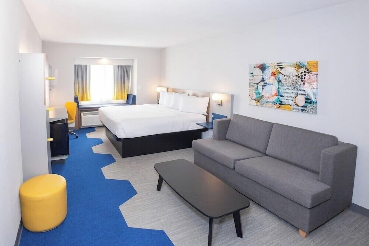 A hotel room with a bed, a sofa, a small table, a yellow stool, and a painting on the wall.