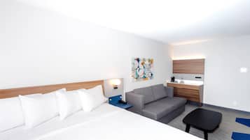 Una camera d'albergo con un letto, un divano grigio, un tavolino, un comodino, una TV a parete e un bagno con lavandino e specchio.