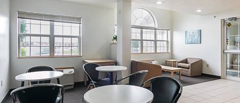 A modern, well-lit room with multiple seating options including round tables and chairs, large windows offering views, and a clean, minimalist design.