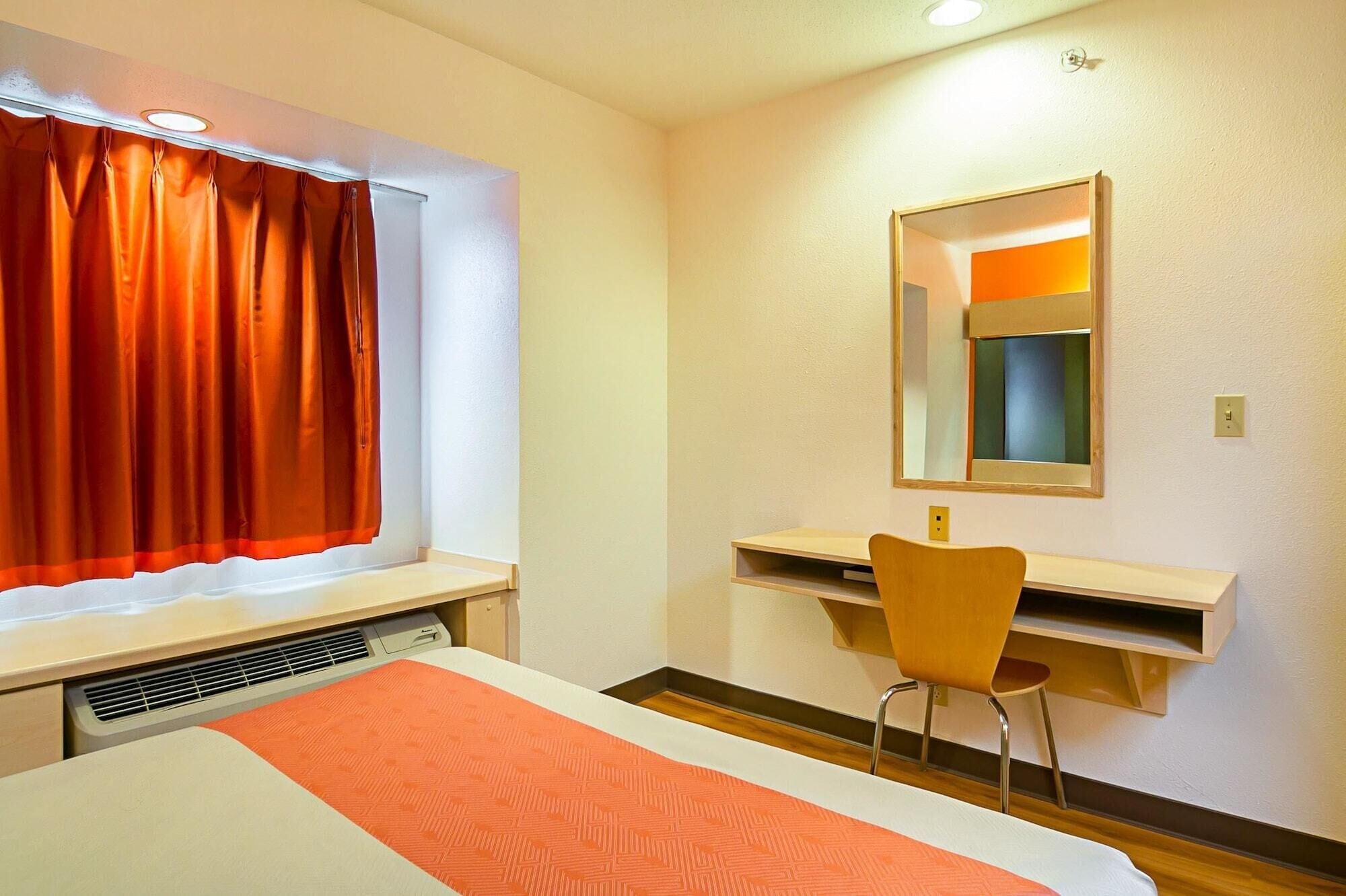 Quarto de hotel com cama, escrivaninha e cadeira, janela com cortinas laranja e espelho na parede.