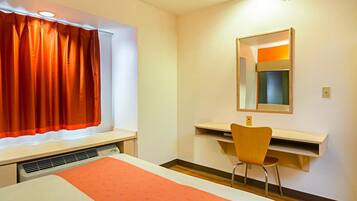 Quarto de hotel com cama, escrivaninha e cadeira, janela com cortinas laranja e espelho na parede.