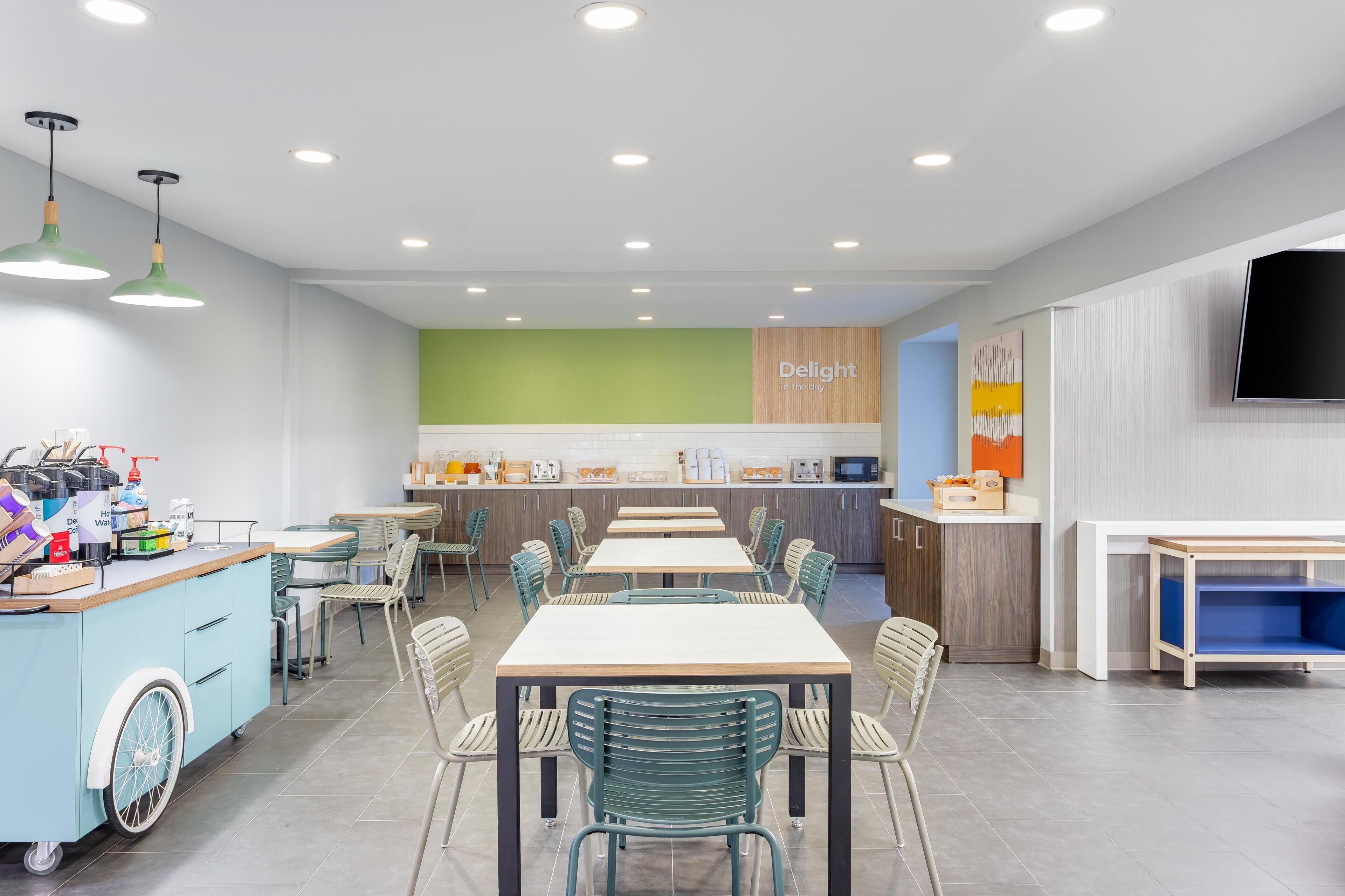 A modern cafeteria with multiple tables and chairs, a counter with food items, and a television mounted on the wall.