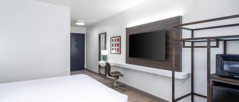 A hotel room with a bed, desk, chair, TV, and a minibar.