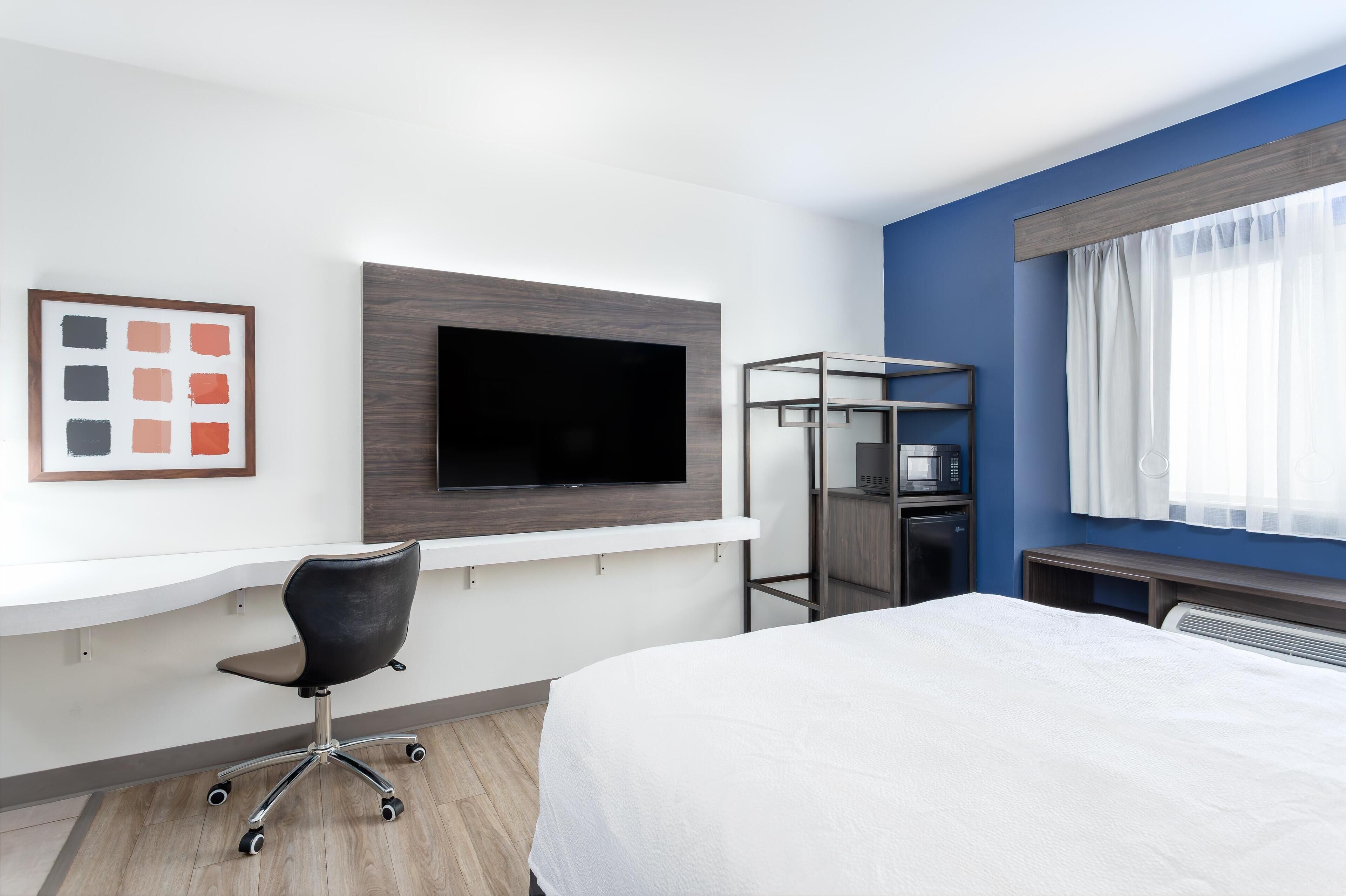 A hotel room with a bed, a desk with a chair, a flat-screen TV mounted on the wall, a microwave, and a small shelf.