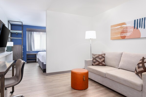 Una habitación de hotel moderna con una cama, un sofá, un escritorio con una silla, un pequeño taburete naranja, un televisor y un cuadro en la pared.