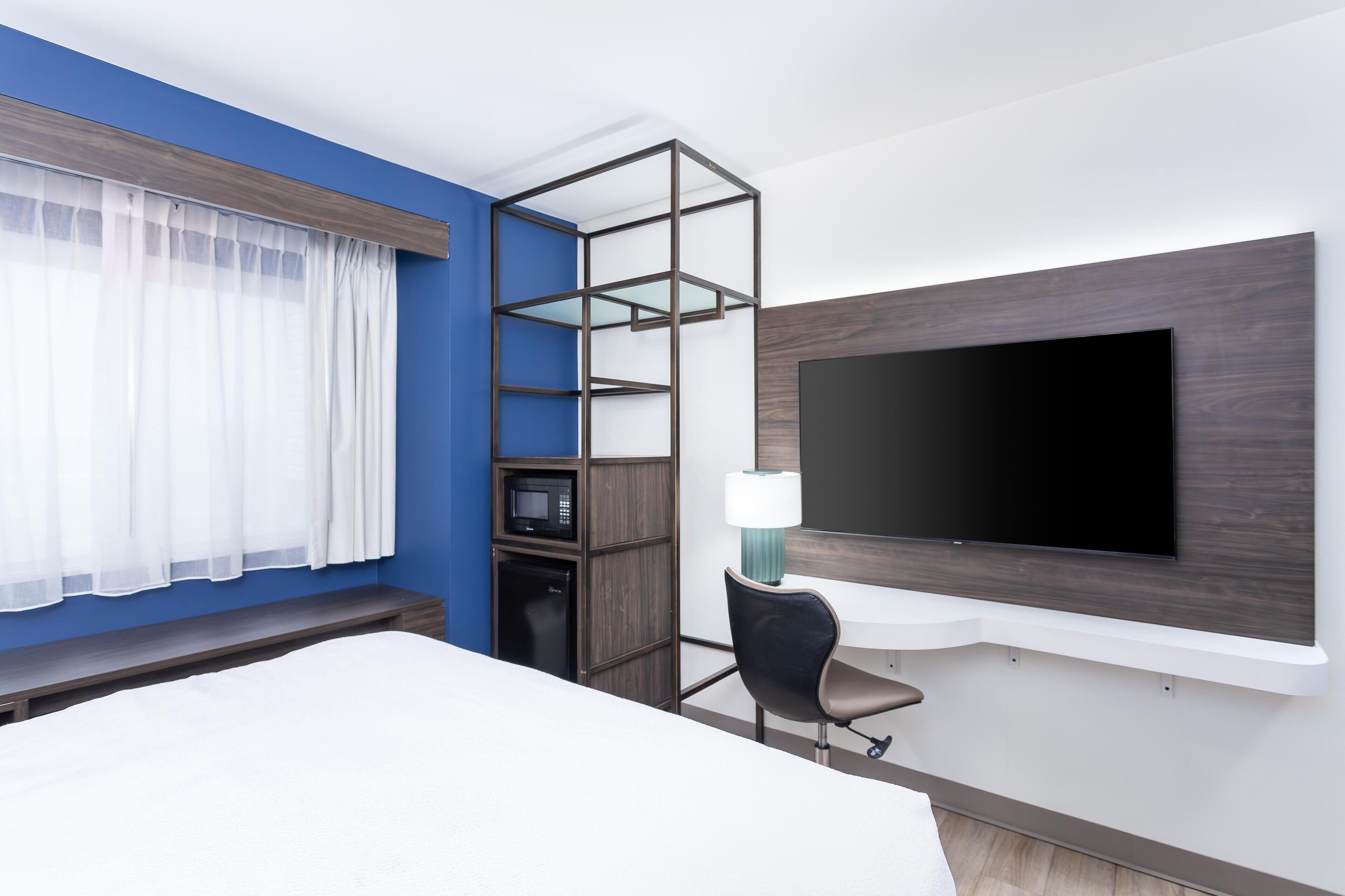 A hotel room with a bed, a TV mounted on the wall, a microwave, a small cabinet, a chair, and a window with sheer curtains.