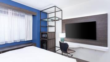 A hotel room with a bed, a TV mounted on the wall, a microwave, a small cabinet, a chair, and a window with sheer curtains.