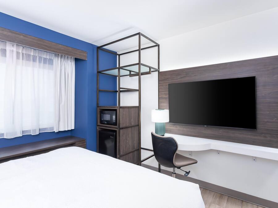 A hotel room with a bed, a TV mounted on the wall, a microwave, a small cabinet, a chair, and a window with sheer curtains.
