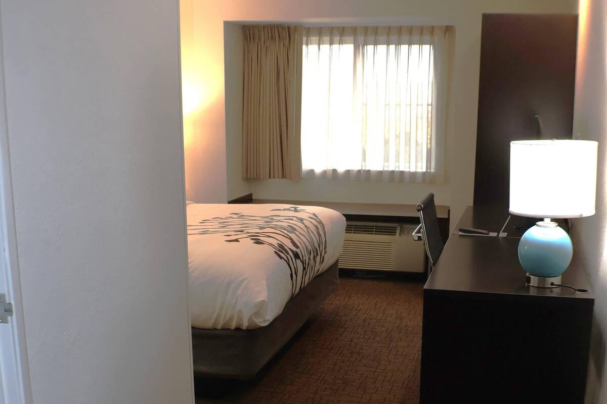 A hotel room with a bed, a desk with a lamp, a window with curtains, and an air conditioning unit.