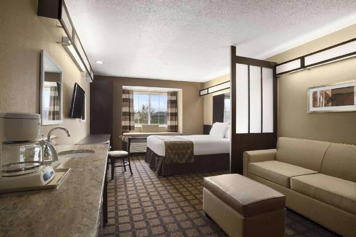A hotel room with a bed, a sofa, a small table with a kettle, a chair, a window with curtains, and a flat-screen TV mounted on the wall.