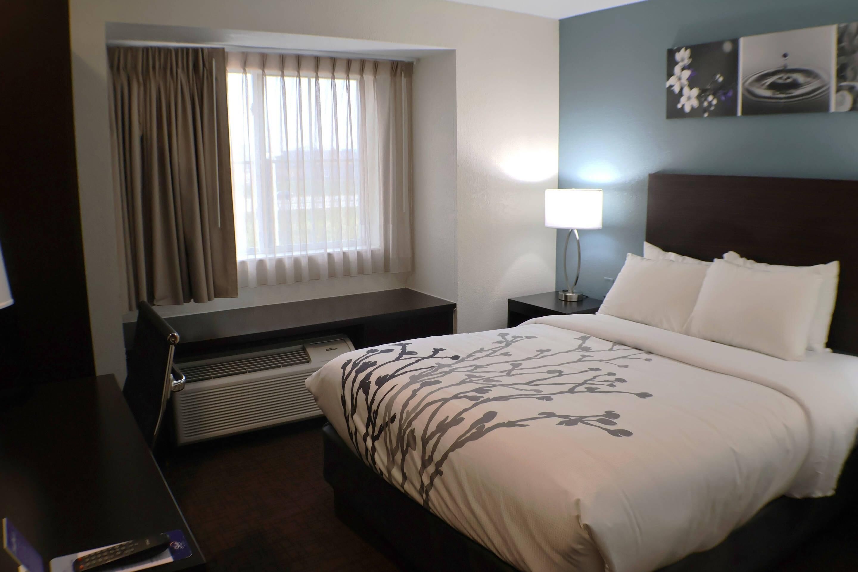 Una habitación de hotel con cama, escritorio, silla, ventana con cortinas, lámpara de pared y radiador.