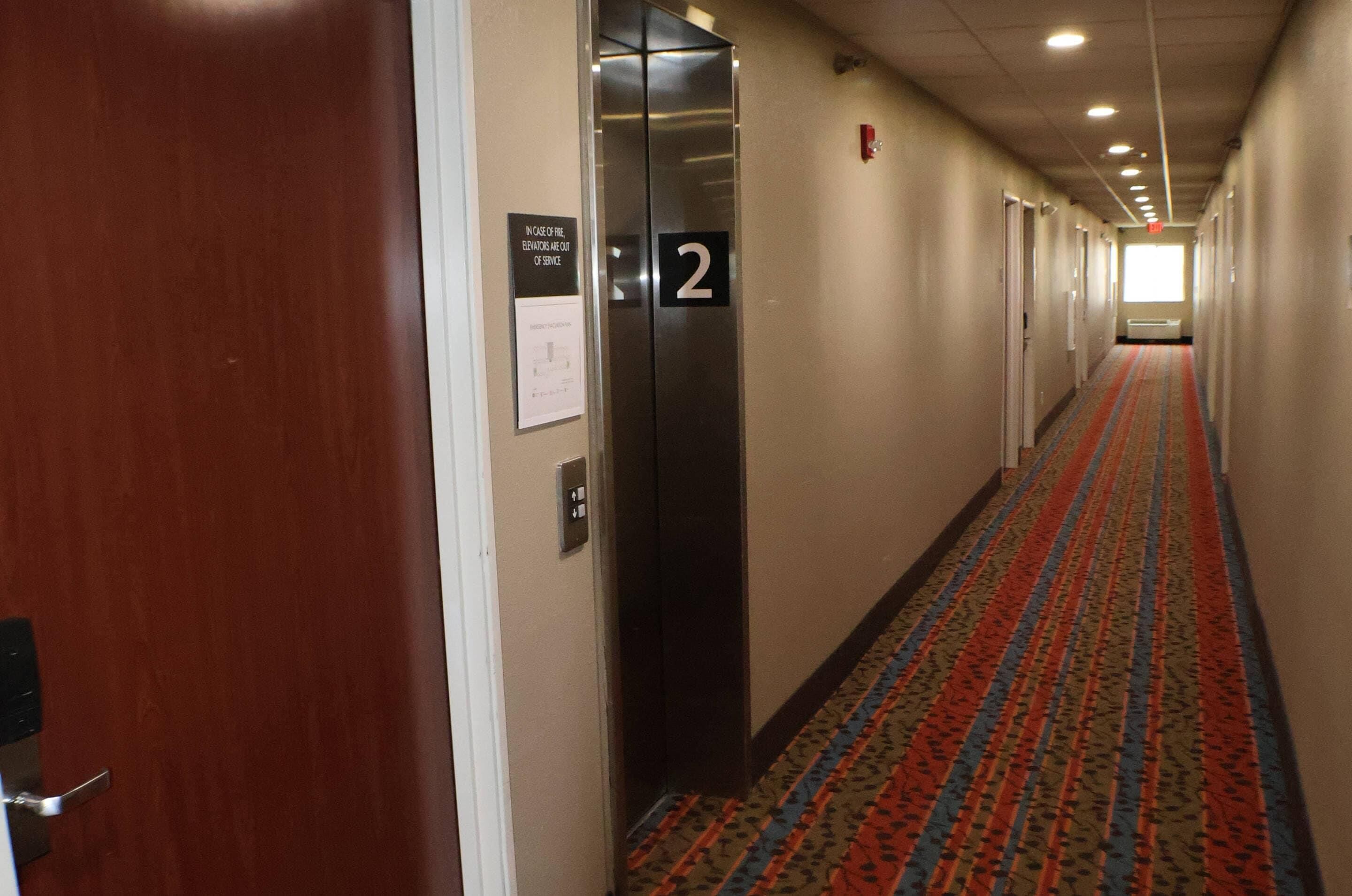 Pasillo de hotel con un ascensor indicado con el número '2', piso alfombrado y puertas a ambos lados.