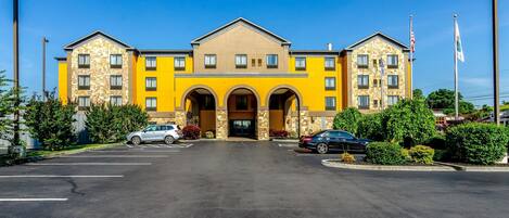 A multi-story hotel with a yellow facade, large entrance with arches, and a parking lot with several cars.