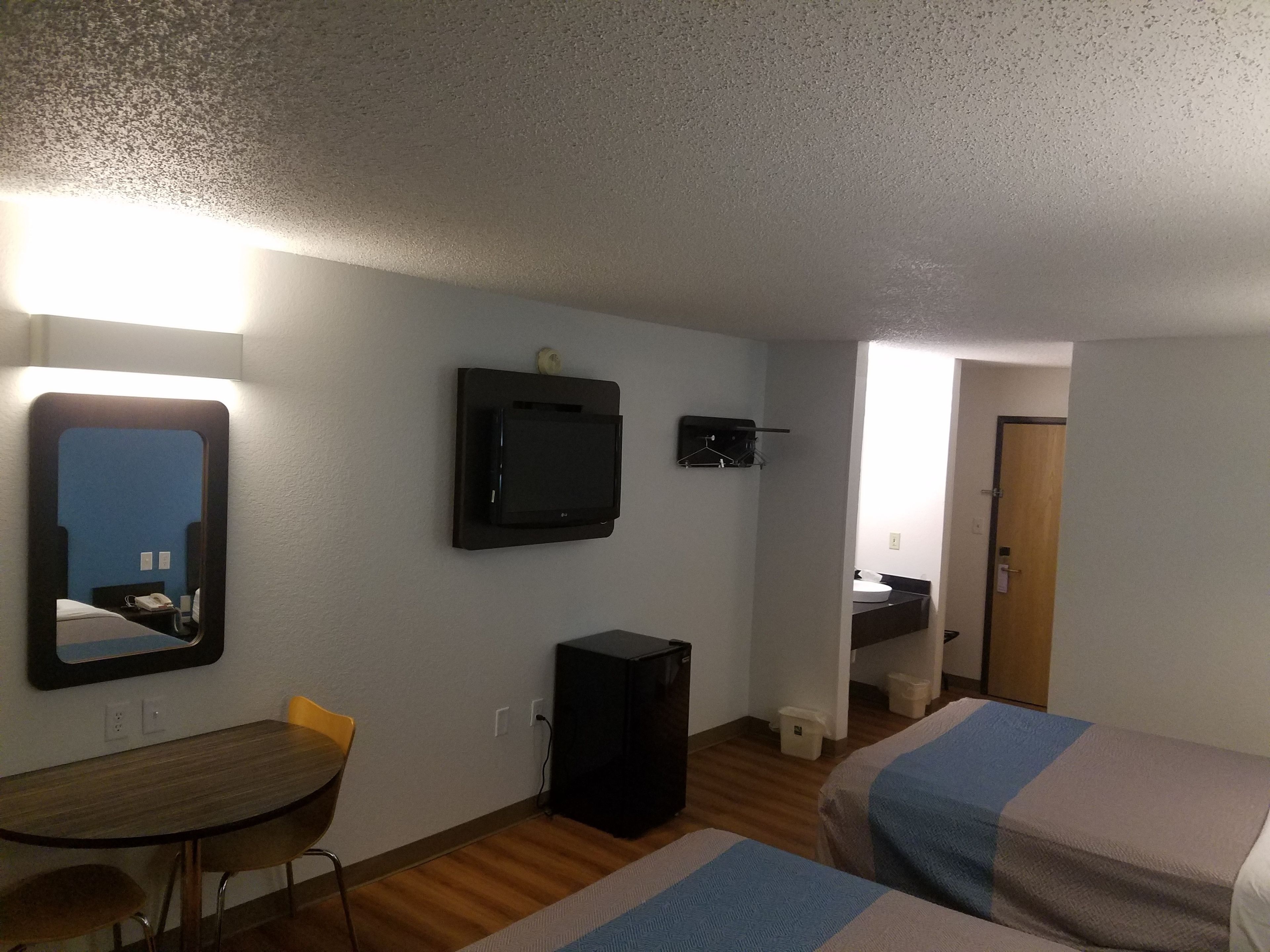 A hotel room with a bed, a desk with a chair, a television mounted on the wall, and a bathroom visible through an open door.