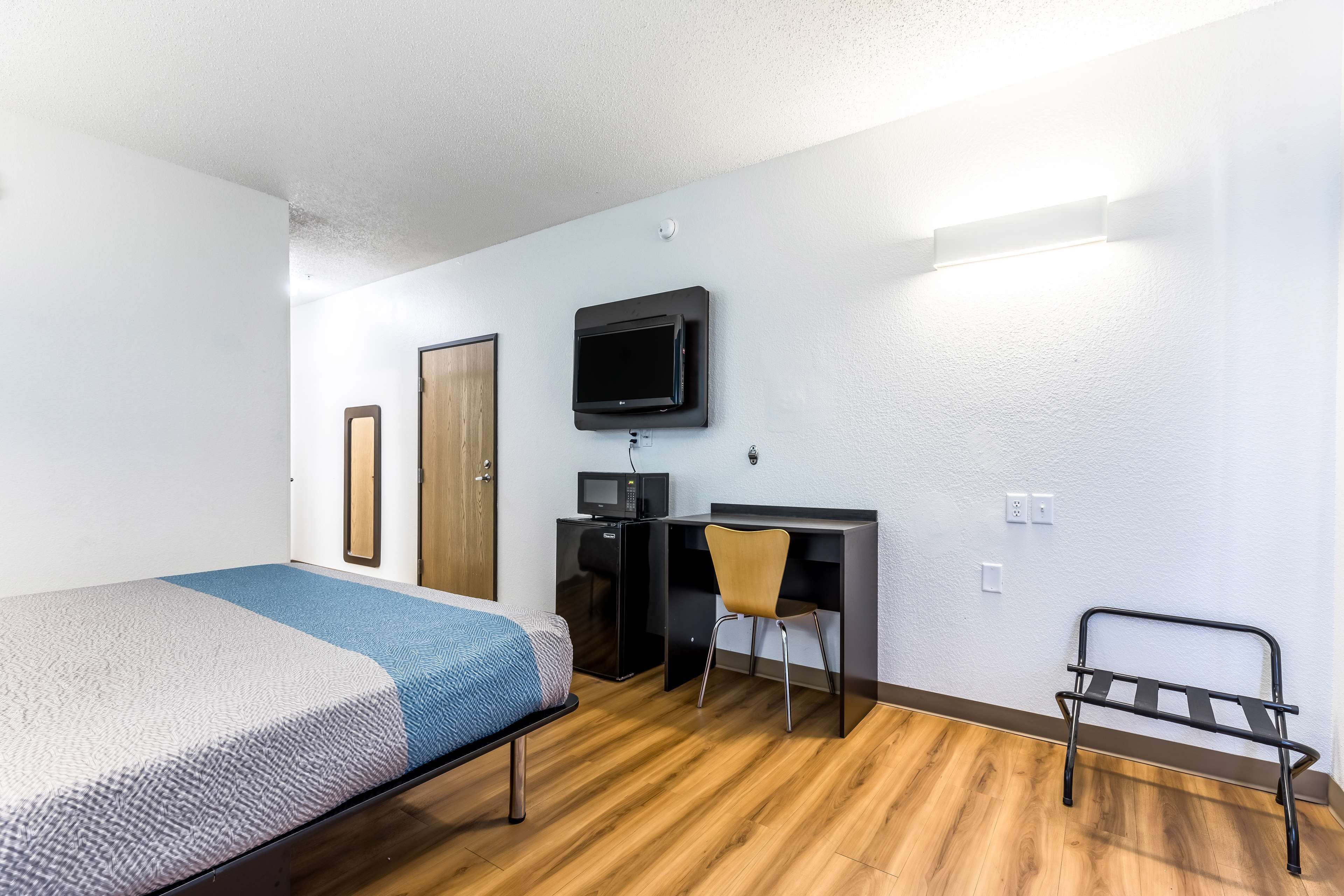 A compact room with a bed, a desk, a chair, a TV mounted on the wall, and a small rack.