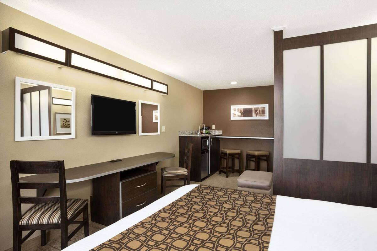 A hotel room with a bed, a desk, a chair, a TV, a kitchen area, and a seating area with stools.