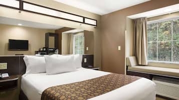 A hotel room with a large bed, a TV, a desk with a phone, a window with curtains, and a view of trees.
