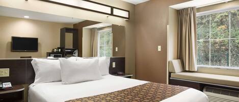 A hotel room with a large bed, a TV, a desk with a phone, a window with curtains, and a view of trees.