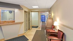 A hotel reception area with a reception desk, a clock, a waiting area with chairs and a table, and a hallway leading to other rooms. - Super 8 by Wyndham Long Island City LGA Hotel (Astoria)