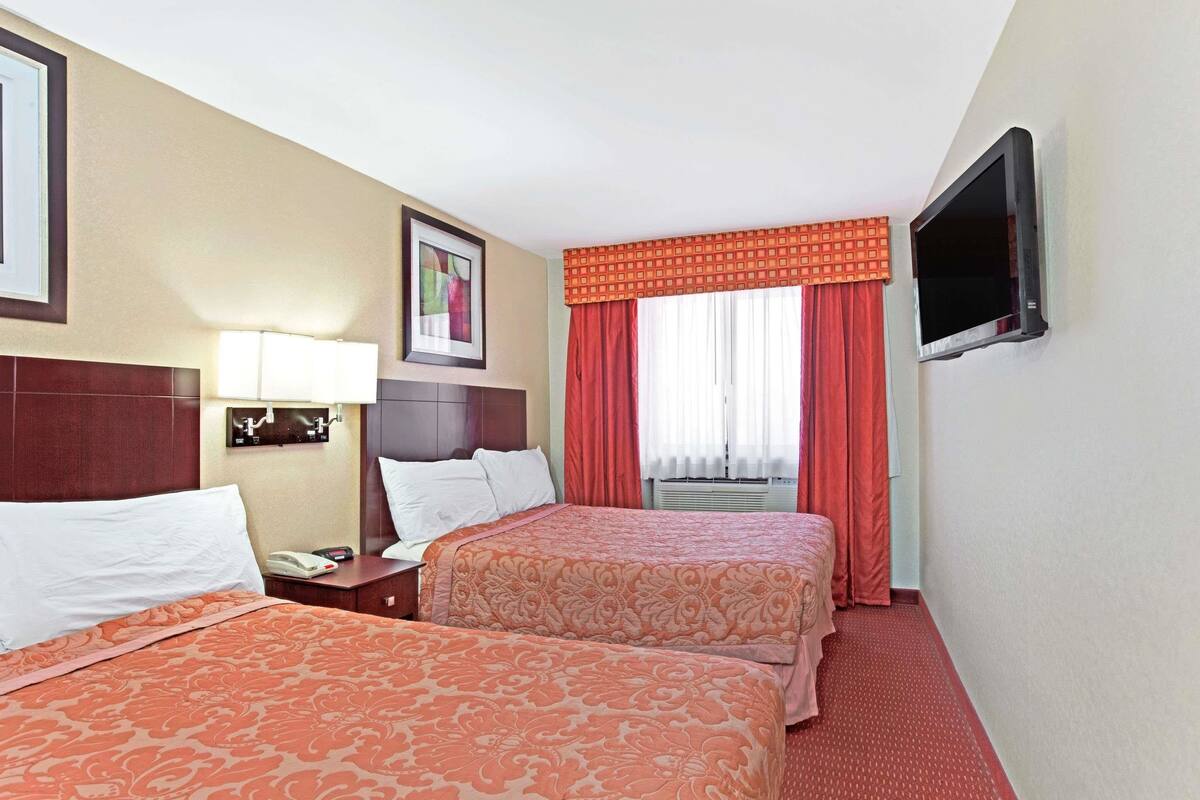 Ein Hotelzimmer mit zwei Betten, einem Fernseher, einem Nachttisch mit Lampe und einem Fenster mit Vorhängen.