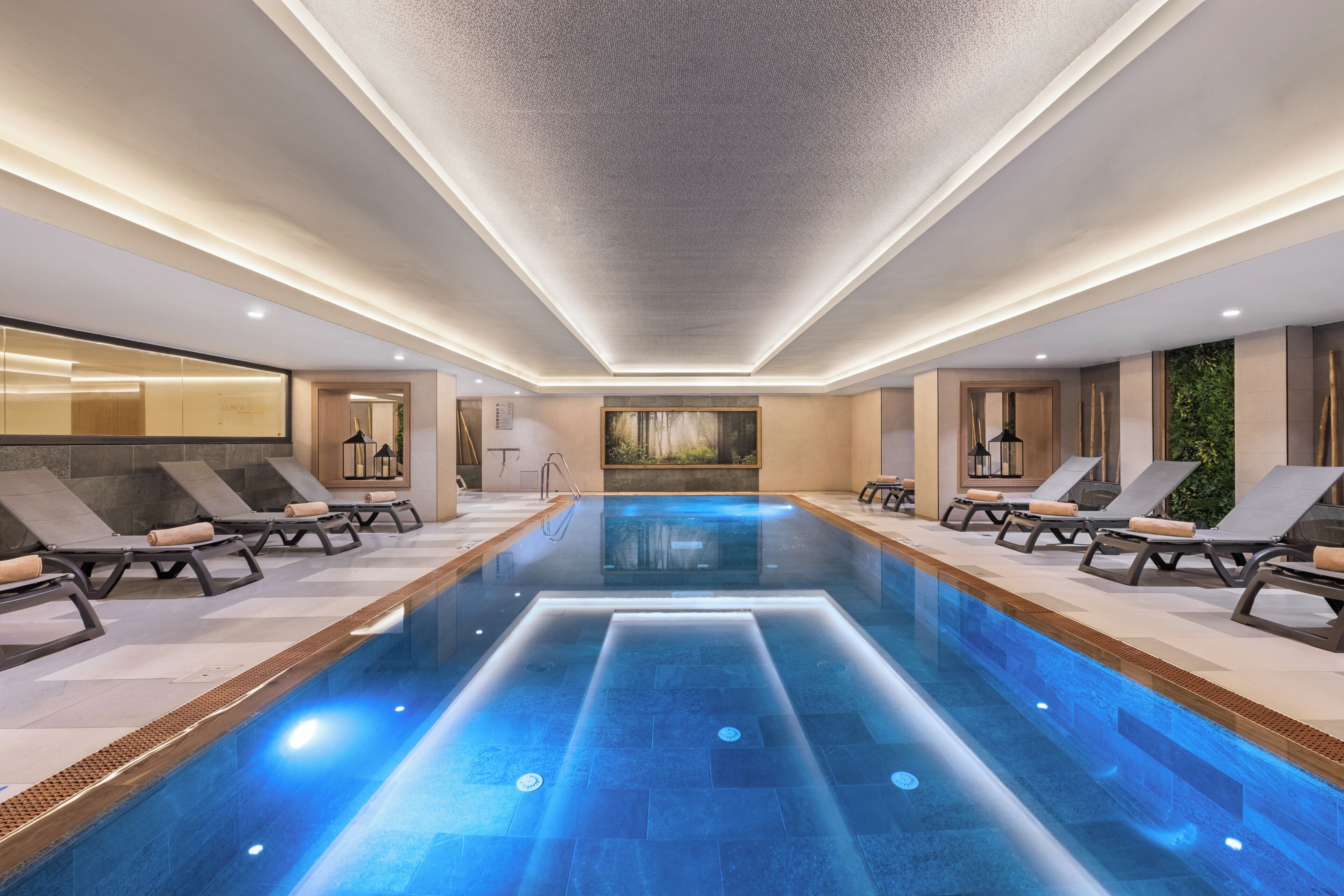 A modern indoor swimming pool with lounge chairs and a view of a forest through a large window.