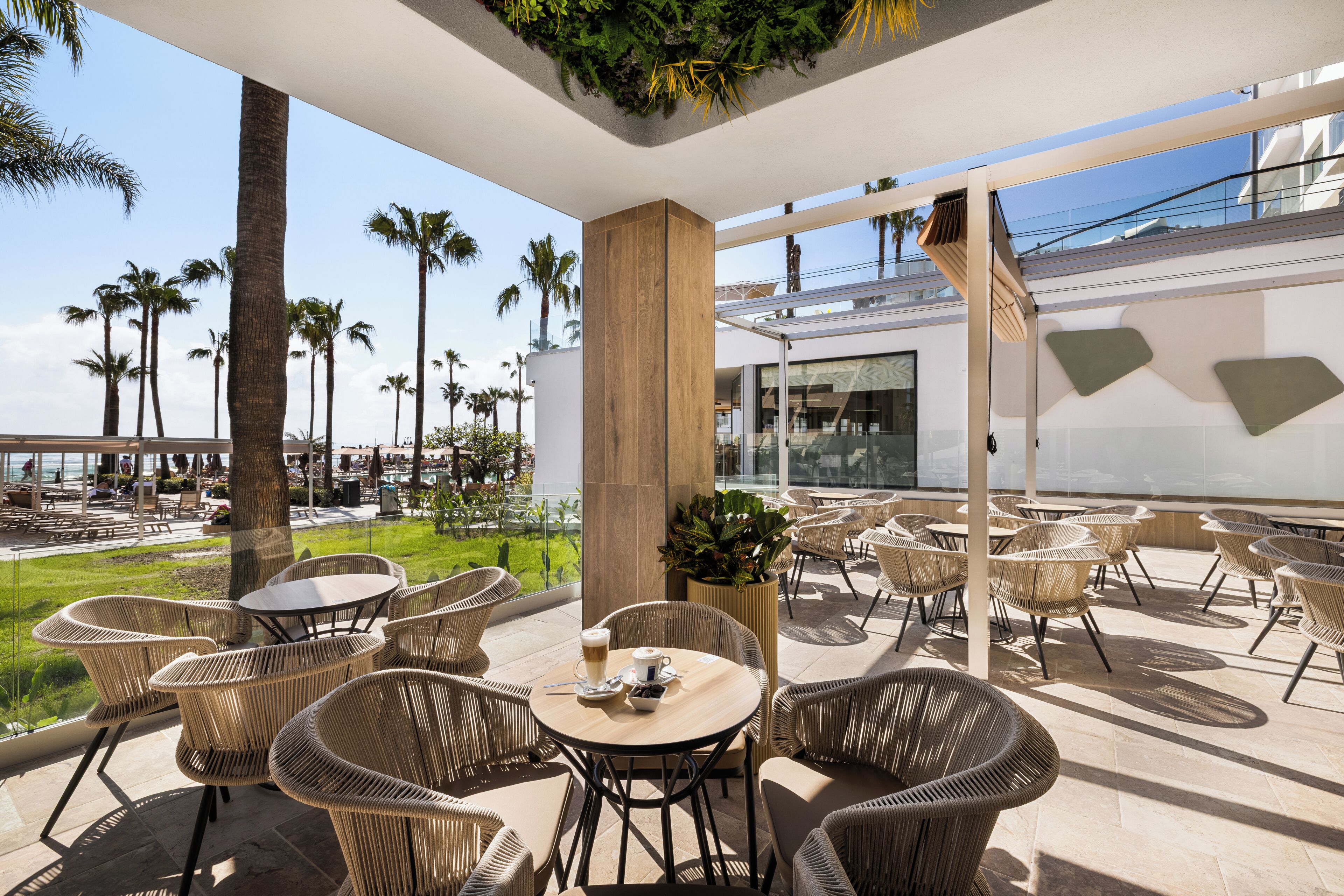 An outdoor patio with wicker furniture, a wooden table, and a view of palm trees and a building in the background.