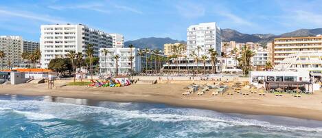 A beachside hotel with multiple floors, palm trees, and a view of mountains in the background.