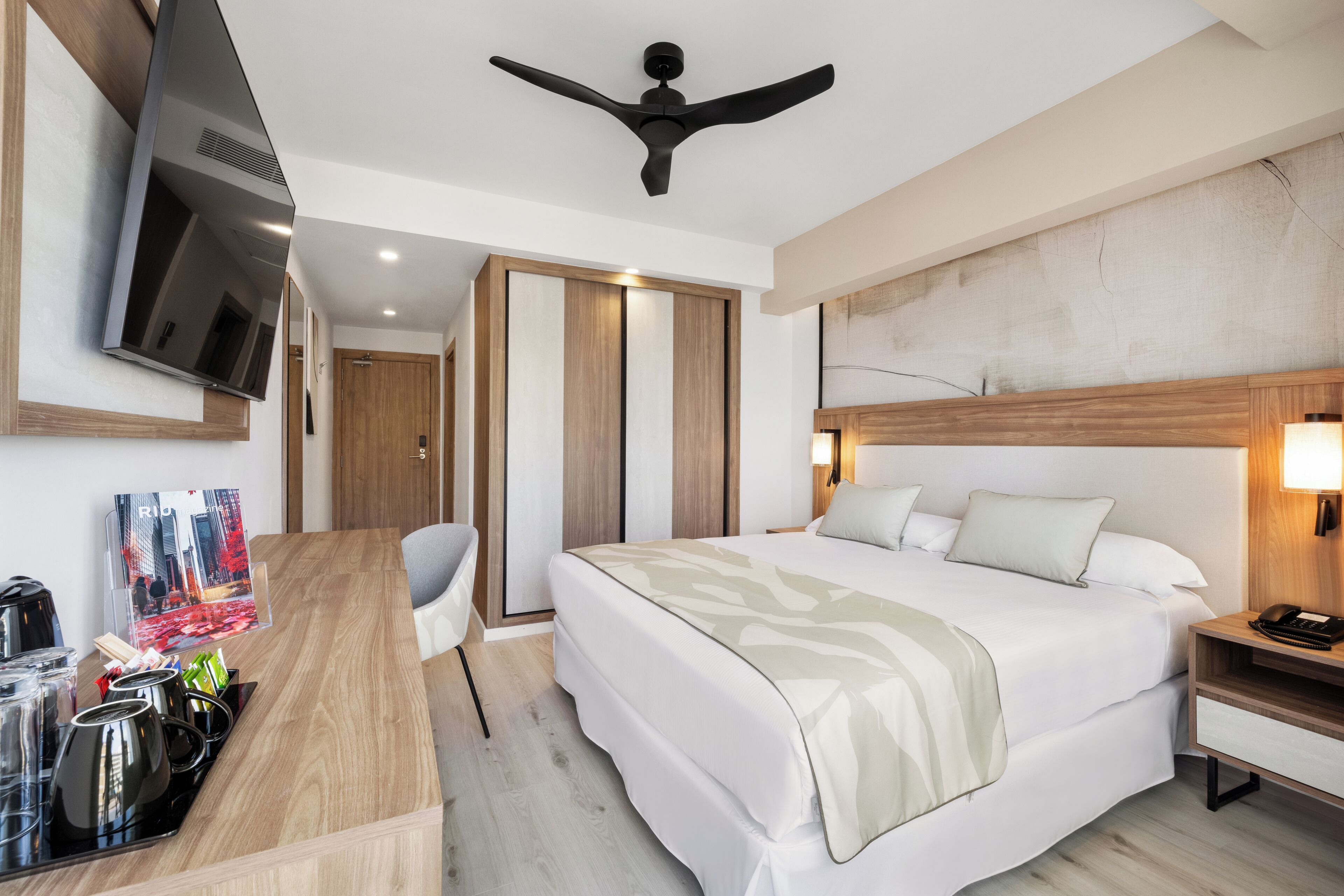 A modern hotel room with a bed, bedside table, TV, ceiling fan, and a desk with coffee-making equipment.