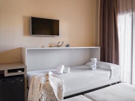 A hotel room with a bed, a television mounted on the wall, a small desk with a refrigerator, and a window with curtains.