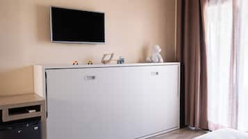 A hotel room with a bed, a TV mounted on the wall, a small desk with a refrigerator, and a window with curtains.