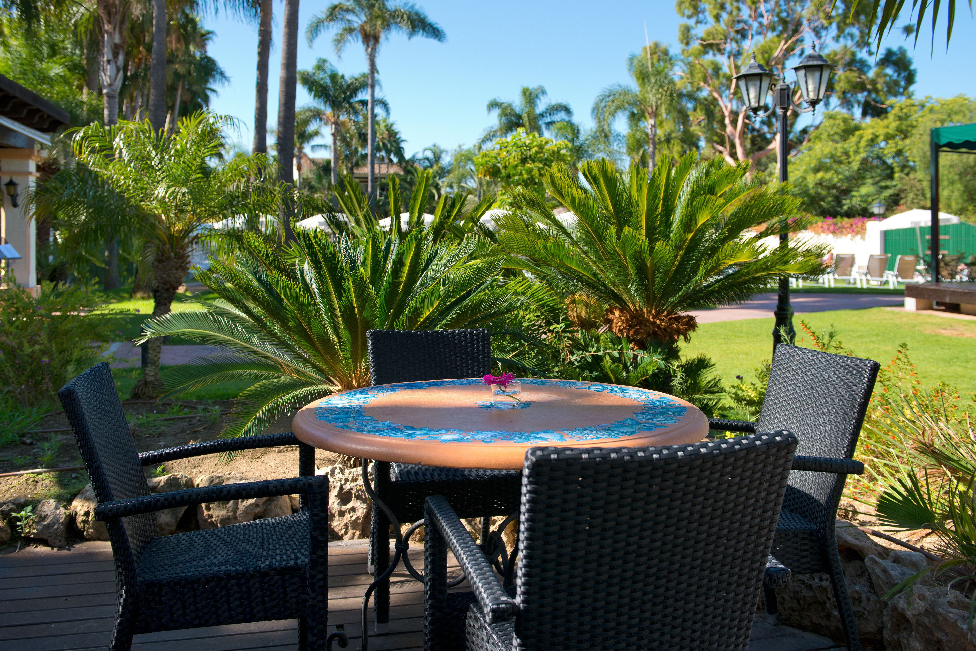 An outdoor dining area with a round table and four chairs, surrounded by lush greenery and palm trees.