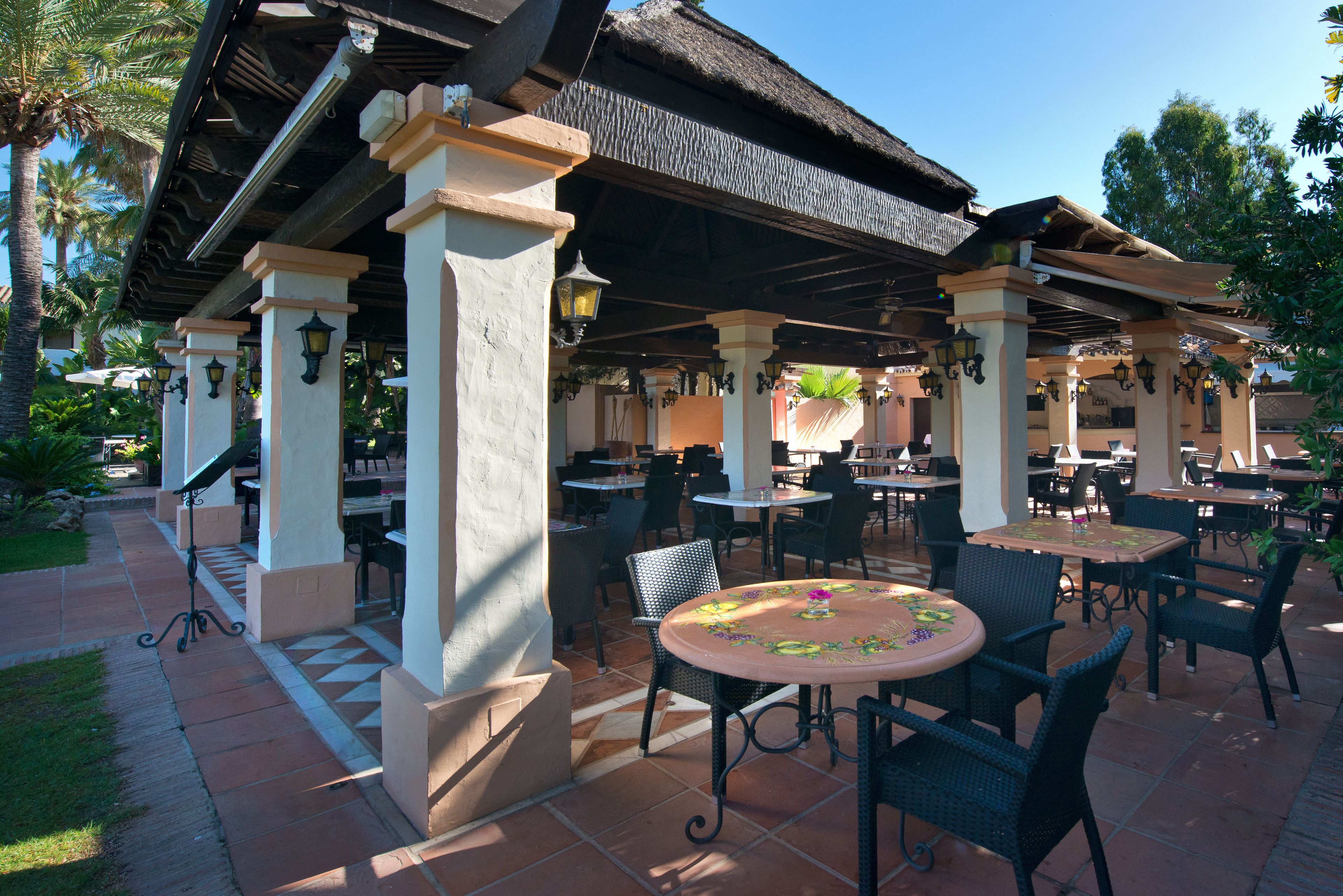 An outdoor patio area with tables and chairs, a thatched roof, and palm trees.