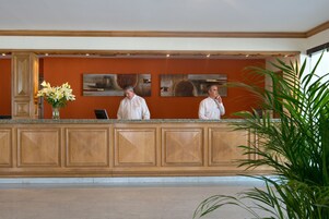 Zona de recepción con un mostrador de madera, dos empleados y un jarrón de flores.
