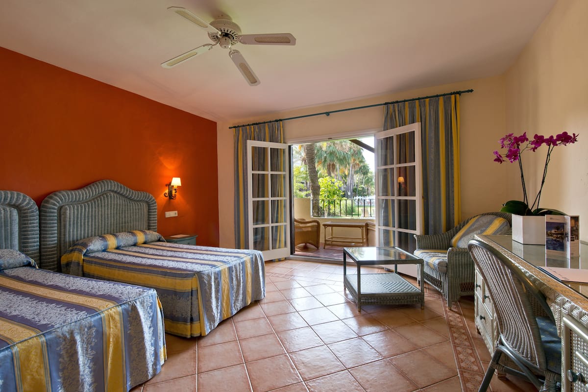 Een hotelkamer met twee bedden, een balkon, een zithoek en een plafondventilator.