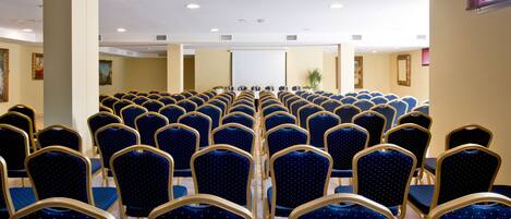 Un salón de conferencias con filas de sillas azules y una pizarra blanca al frente.