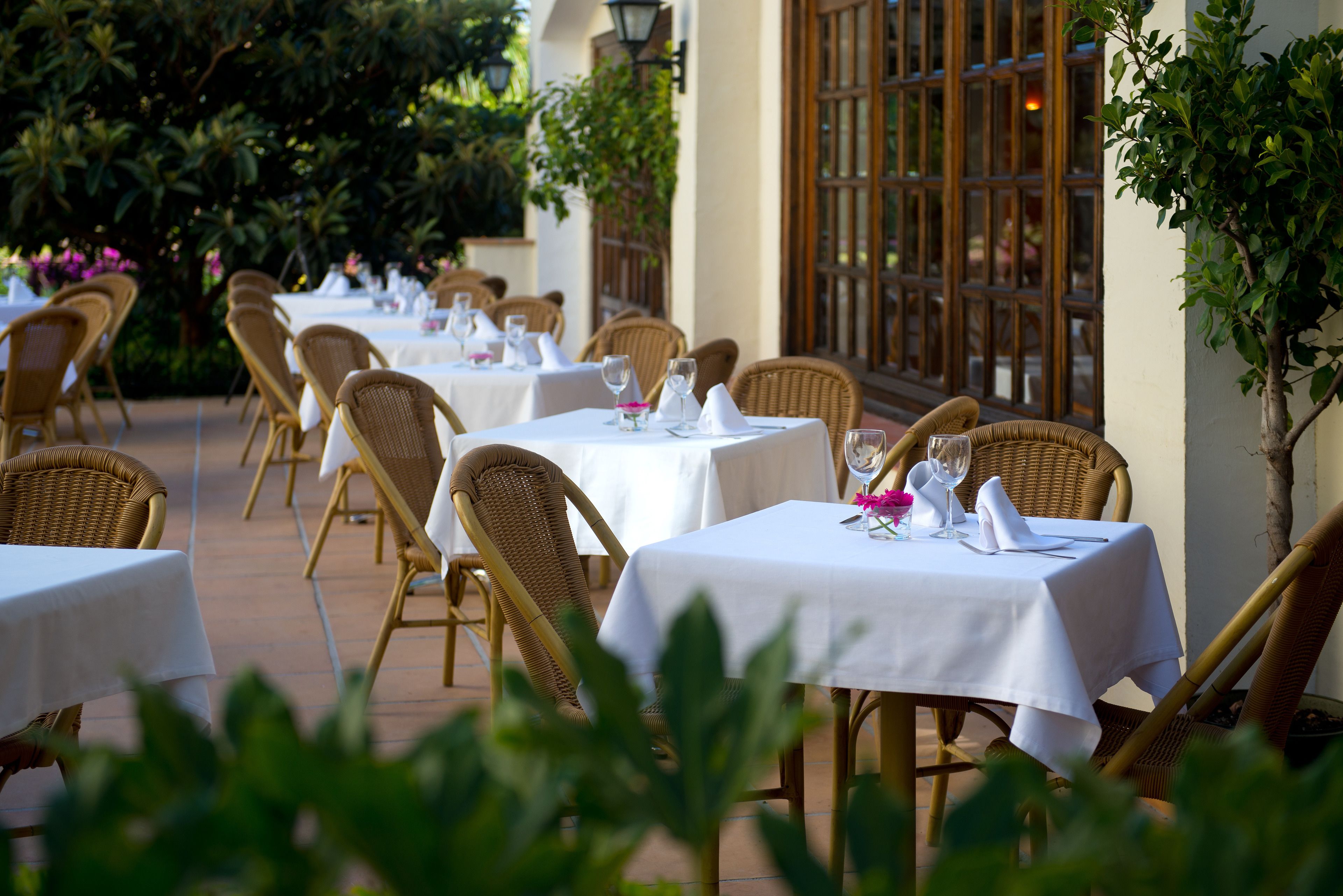 An outdoor dining area with wicker chairs and tables, each set with white tablecloths and glassware.