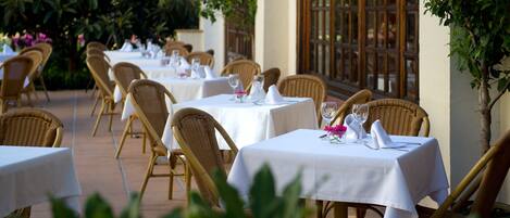An outdoor dining area with wicker chairs and tables, each set with white tablecloths and glassware.