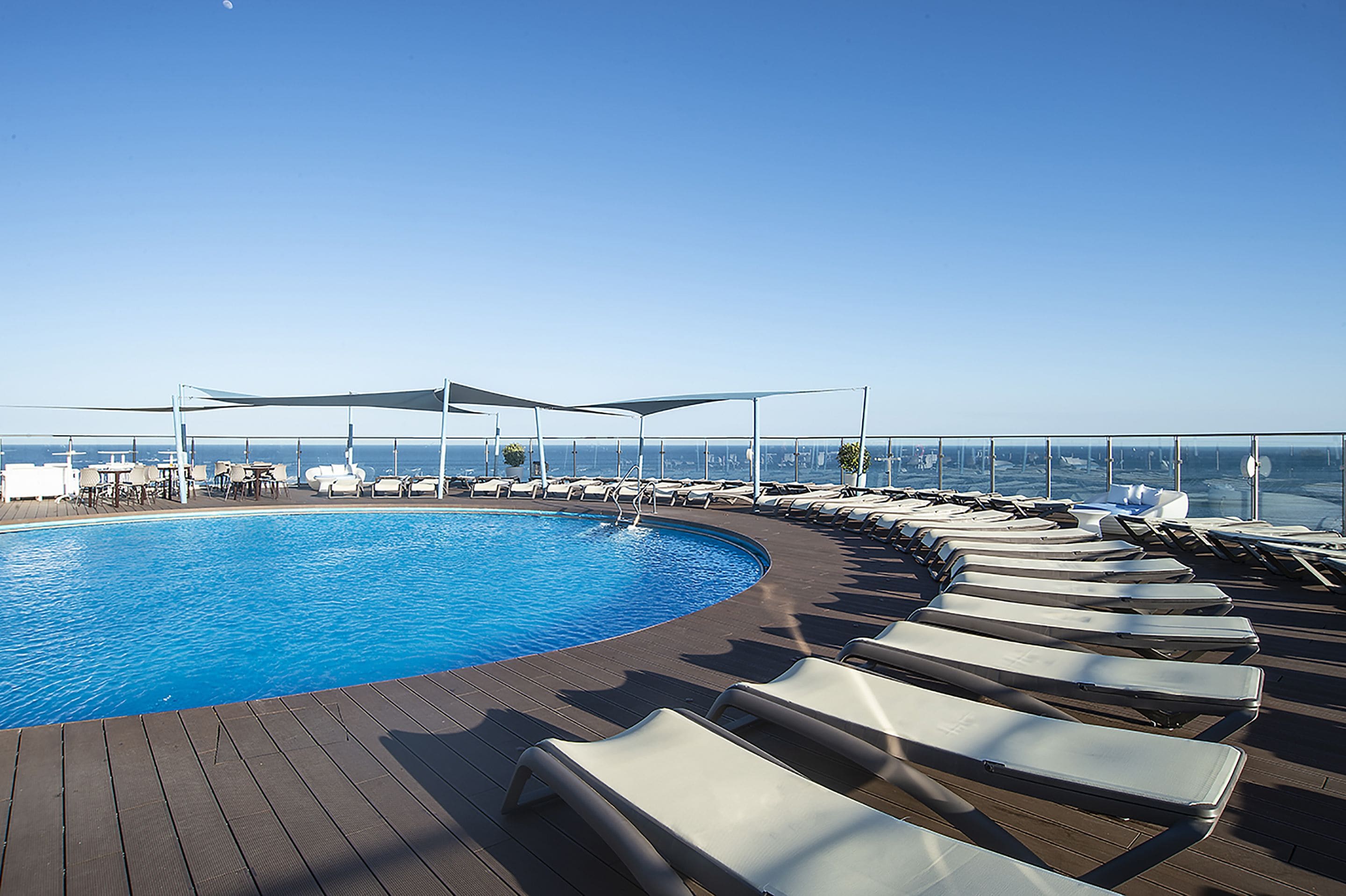 A rooftop pool with sun loungers and umbrellas, surrounded by a wooden deck and overlooking a cityscape.