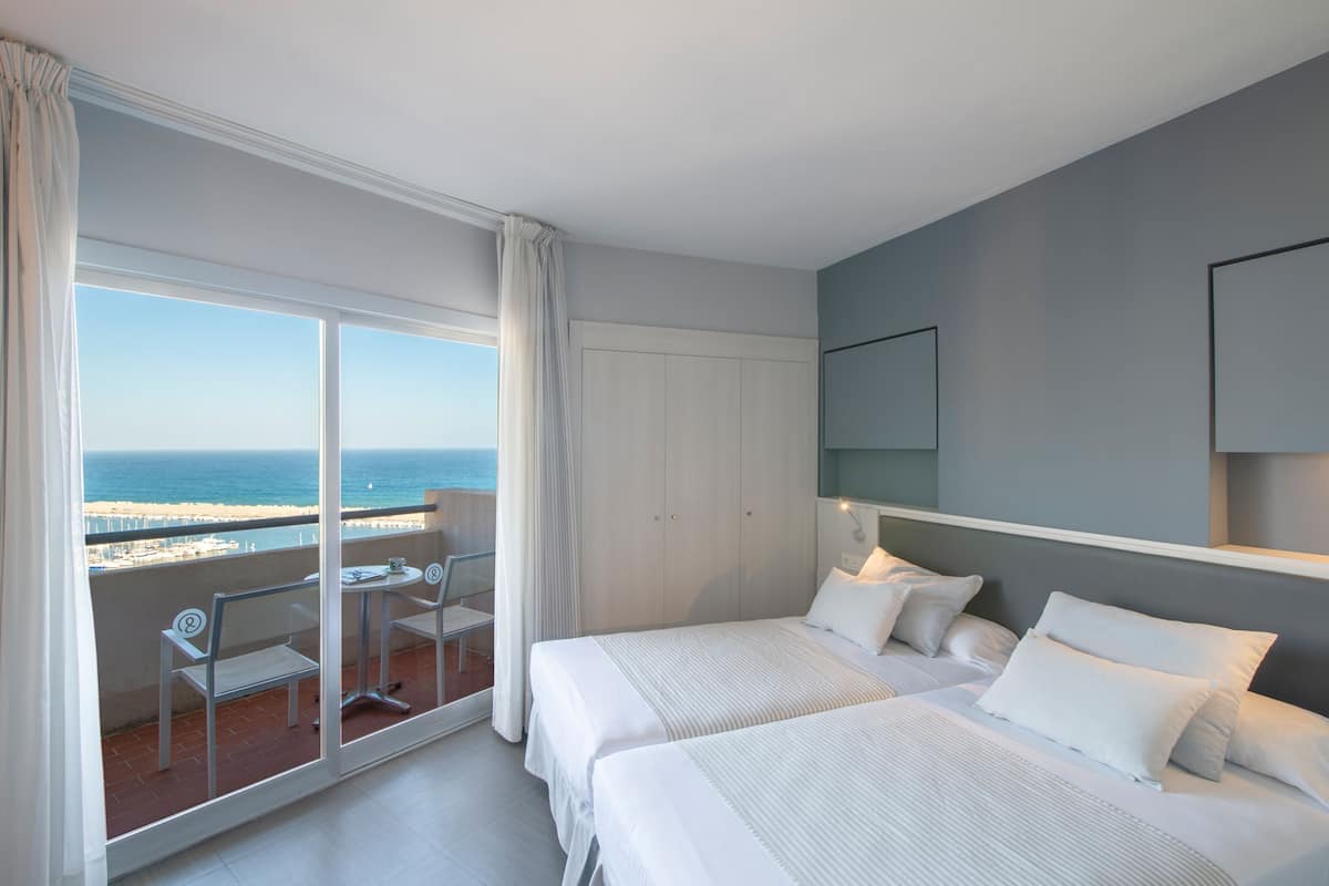 Een hotelkamer met twee bedden, een balkon met uitzicht op zee, en een kleine tafel met stoelen.