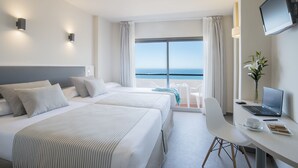 Habitación de hotel con dos camas, un escritorio con una computadora portátil, una silla, una mesita y vista al mar.