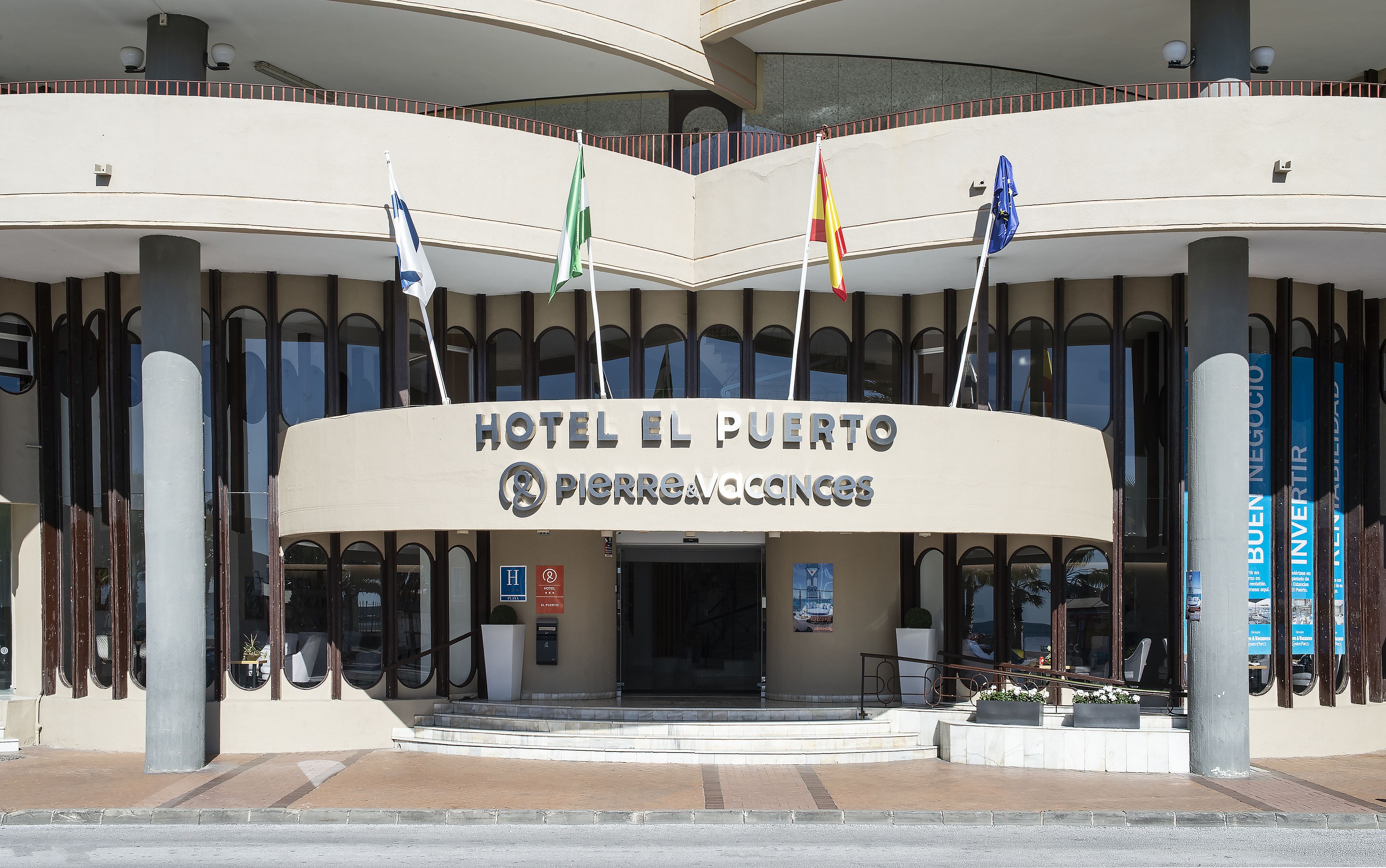 The entrance of HOTEL EL PUERTO with flags, a sign for a parking area, and a view of the interior.