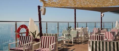 A deck with striped chairs and tables, a lifebuoy, and a view of the water.