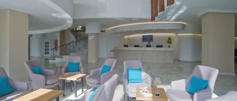 A hotel lobby with a reception desk, seating area with tables and chairs, and a staircase leading to an upper level.