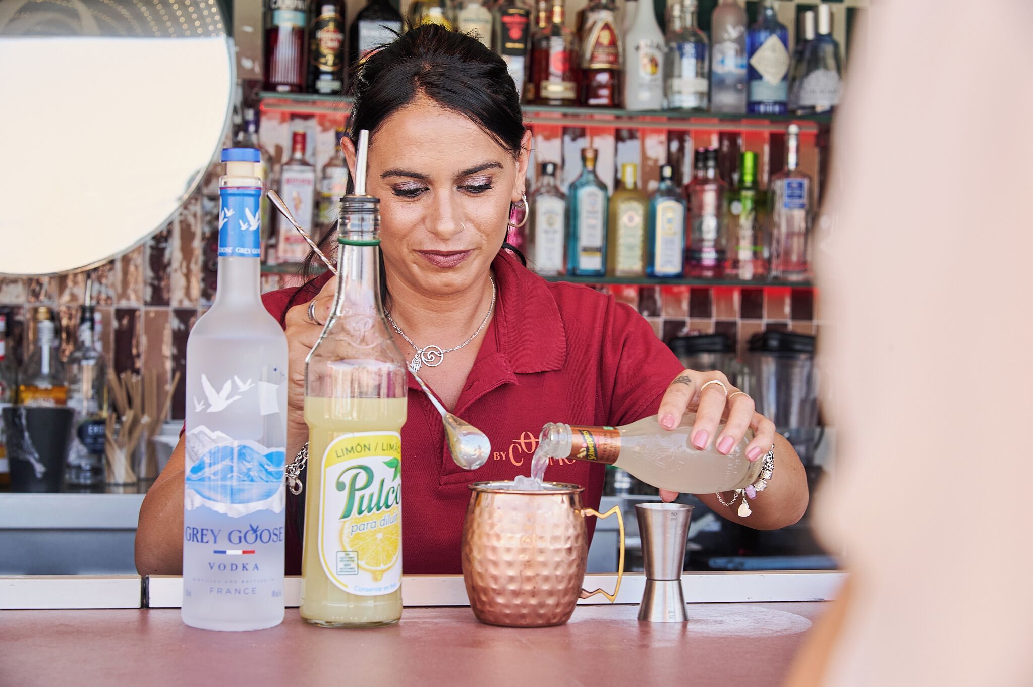 Un barman preparant una beguda amb un got de coure, una ampolla de vodka Grey Goose i una ampolla de Púlver sabor llimona-llauna.
