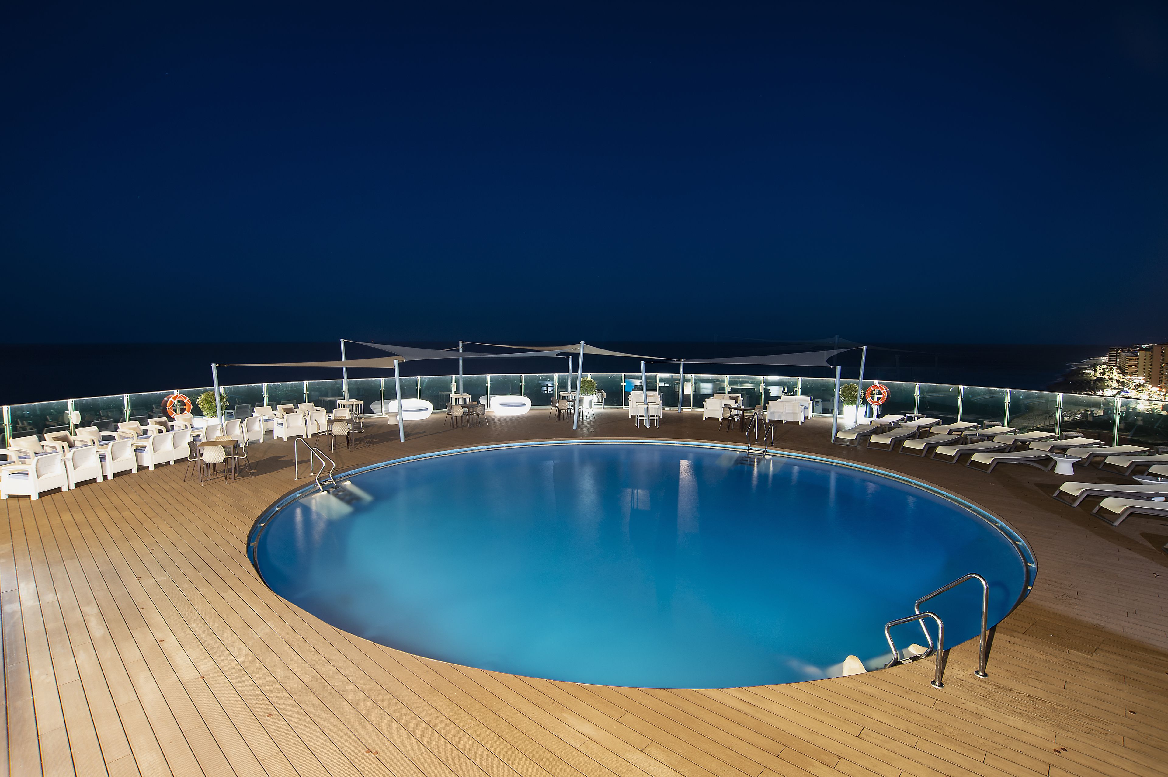 A large, circular pool surrounded by white lounge chairs and tables on a wooden deck, with a clear view of the ocean at night.