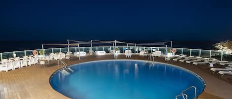 A large, circular pool surrounded by white lounge chairs and tables on a wooden deck, with a clear view of the ocean at night.