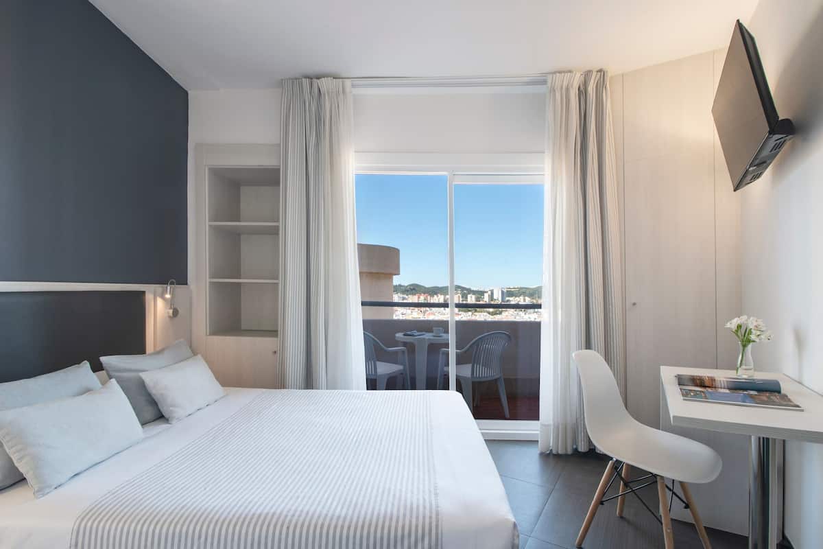 Een hotelkamer met een bed, een bureau en uitzicht op de stad door grote ramen.